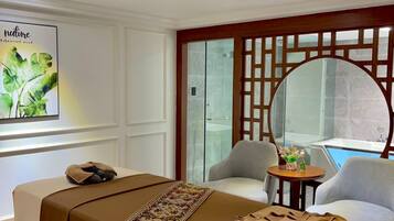 A hotel room with a massage table, two chairs, a small table with flowers, a large mirror, and a poster on the wall.