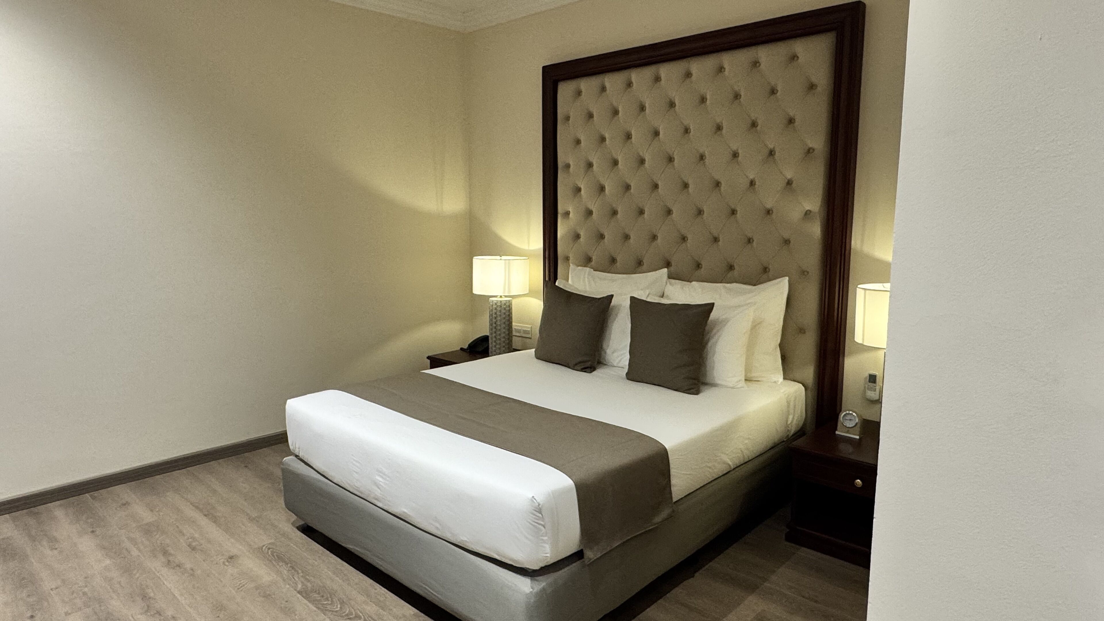 A hotel room with a large bed, a tufted headboard, two bedside lamps, and a nightstand with a phone and a clock.