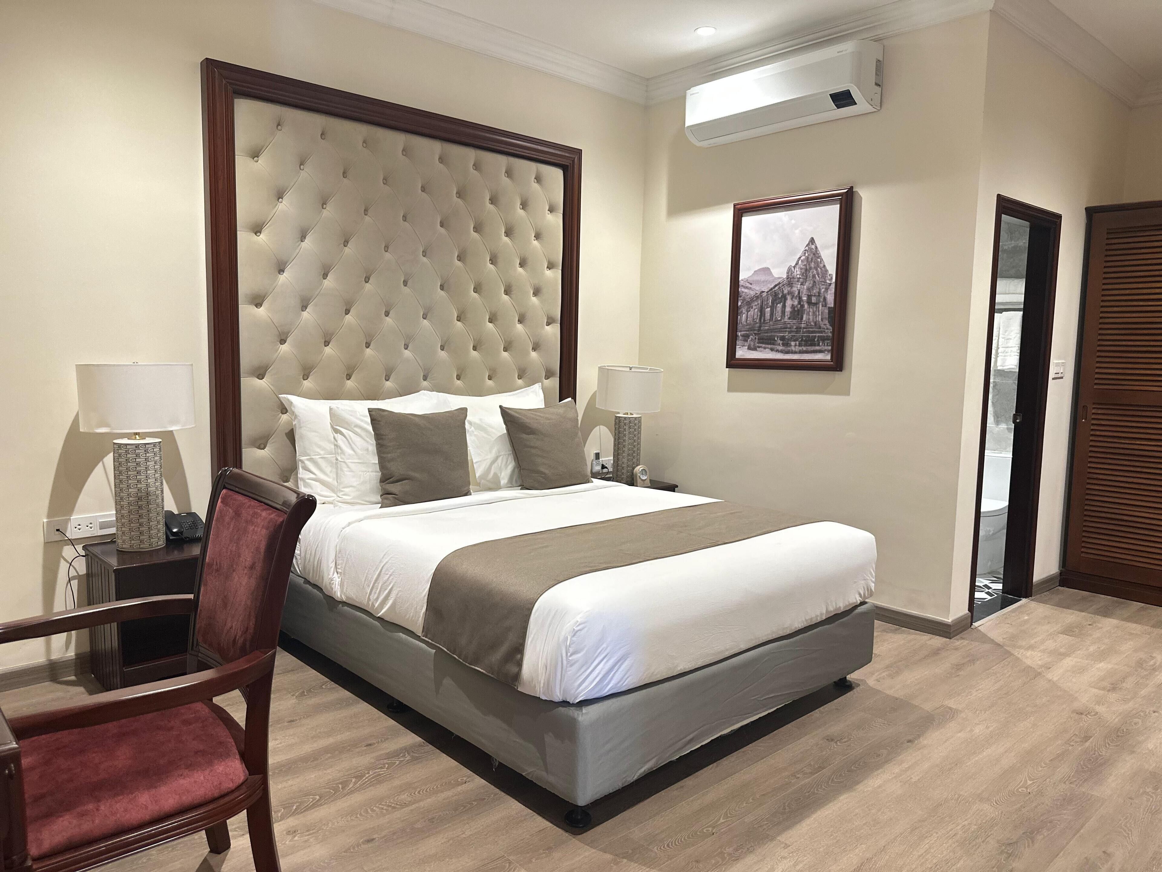 A hotel room with a large bed, two bedside tables, a chair, a wall-mounted air conditioner, and a framed picture.