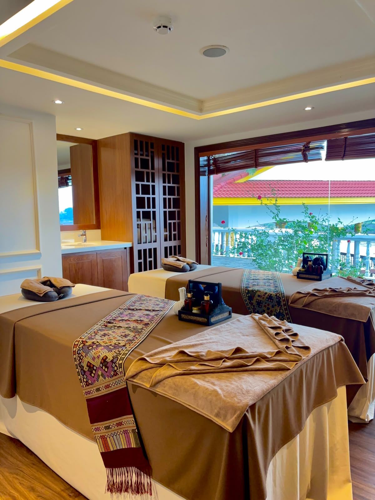 A hotel room with two beds, a sitting area, and a view of a red-roofed building outside.