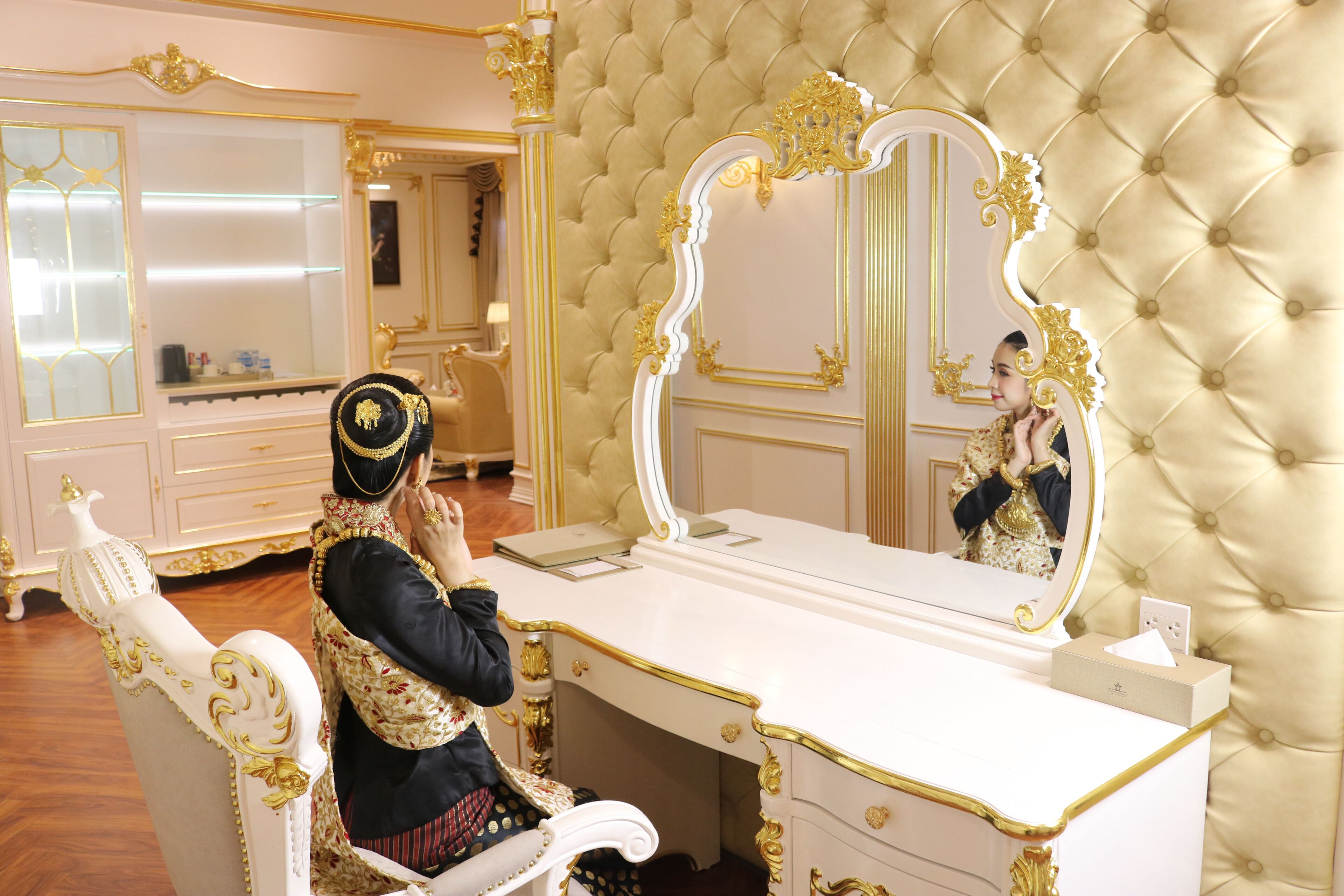 A luxurious room with a tufted wall, a large ornate mirror, and a white vanity with gold accents.