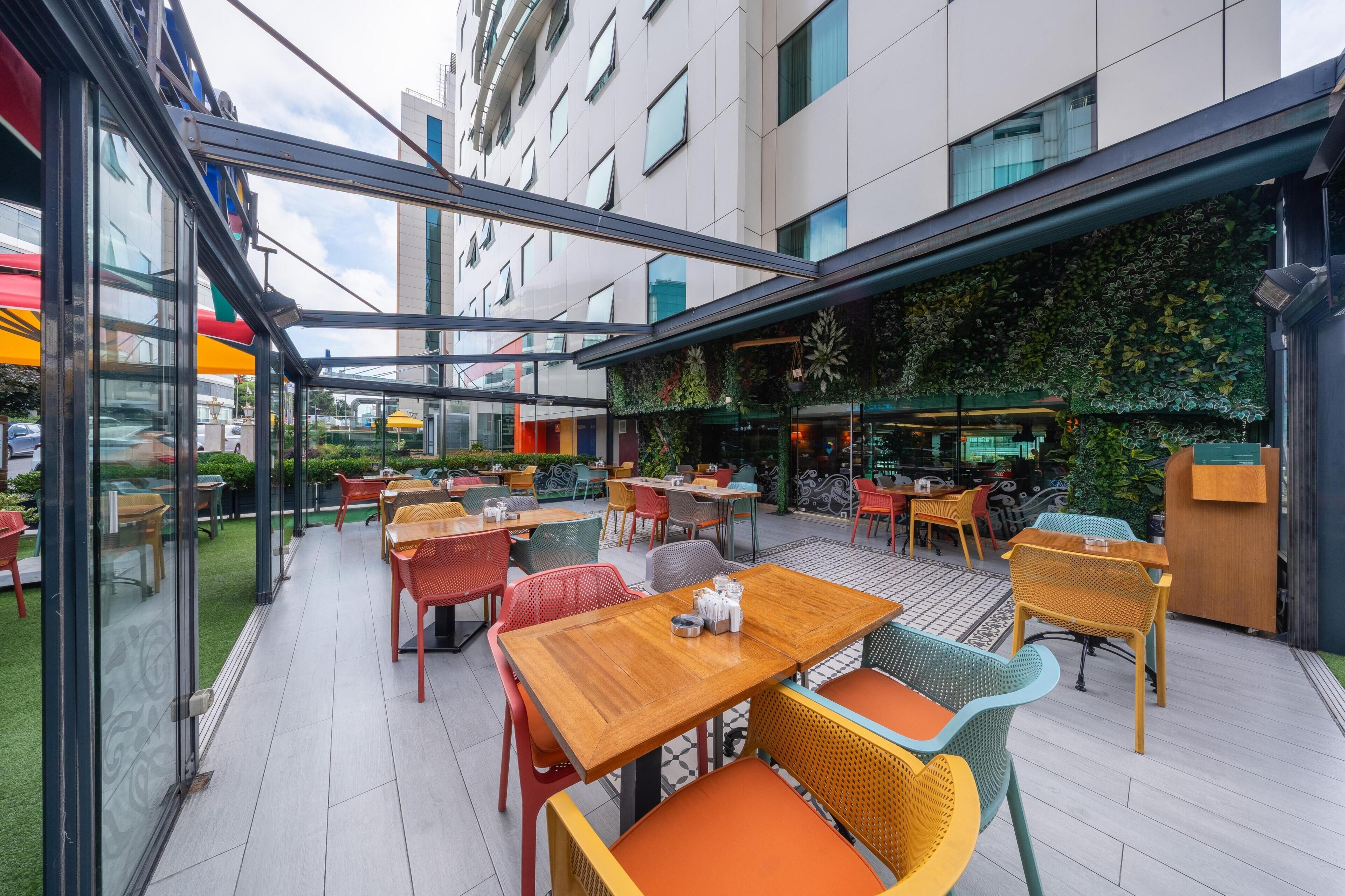 An outdoor cafe area with tables and chairs, a glass enclosure, and a green wall with plants.