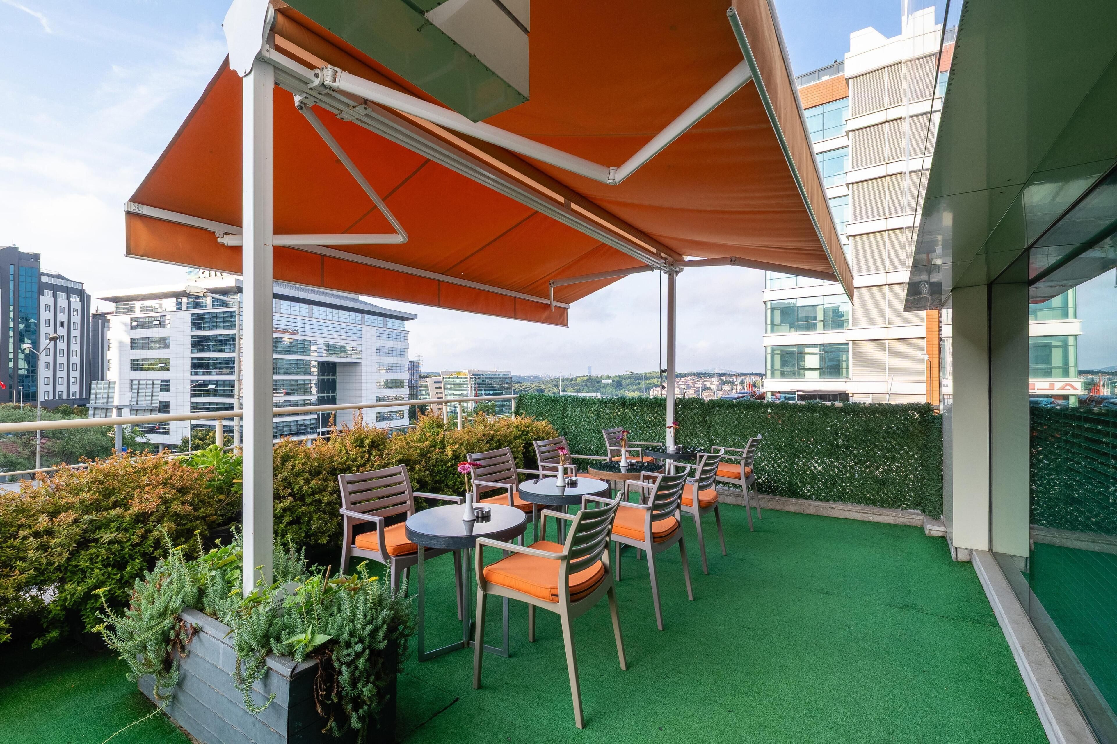 An outdoor seating area with tables and chairs, surrounded by greenery and overlooking a cityscape.