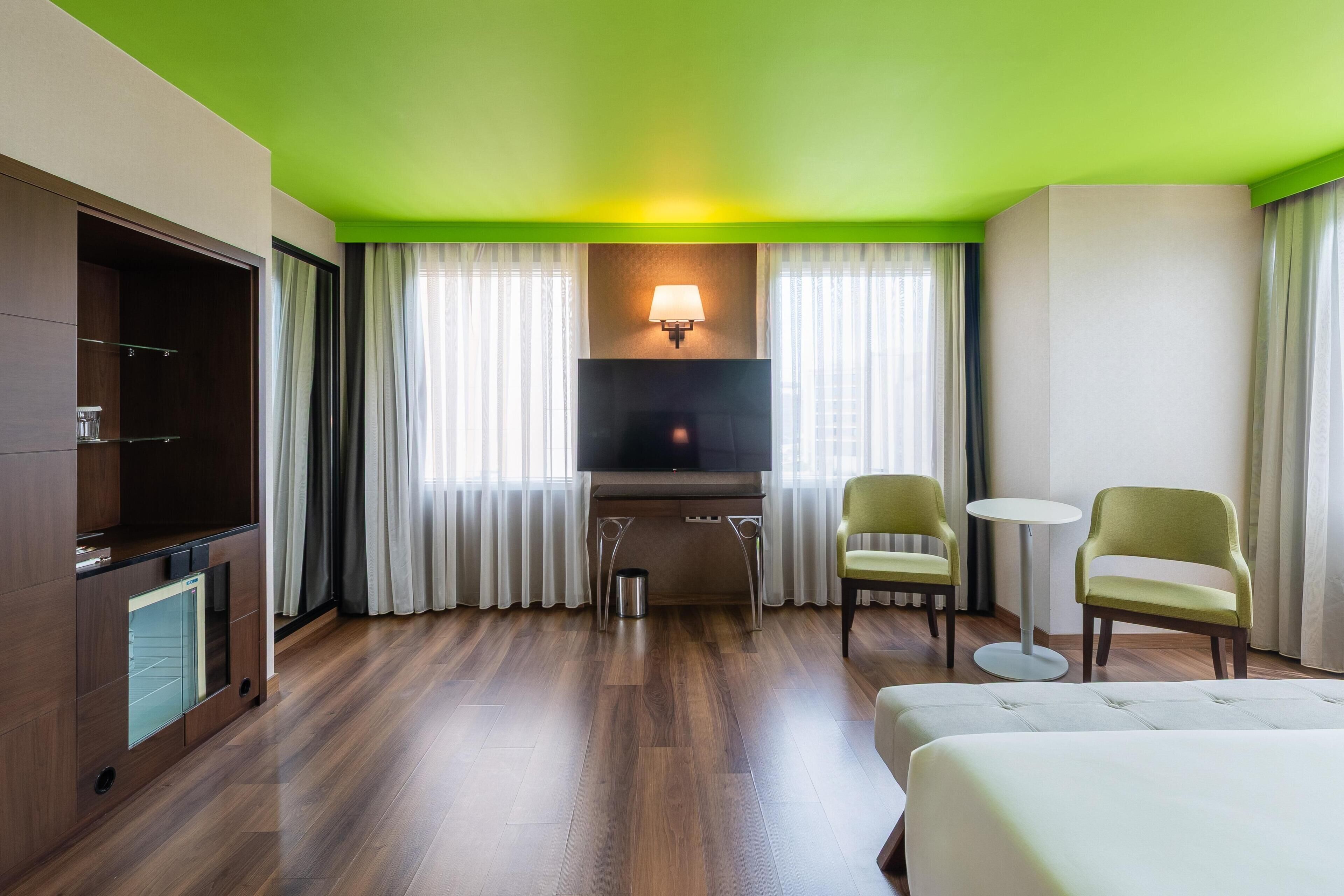 A hotel room with a flat-screen TV, a sofa, two chairs, a small table, and a window with curtains.