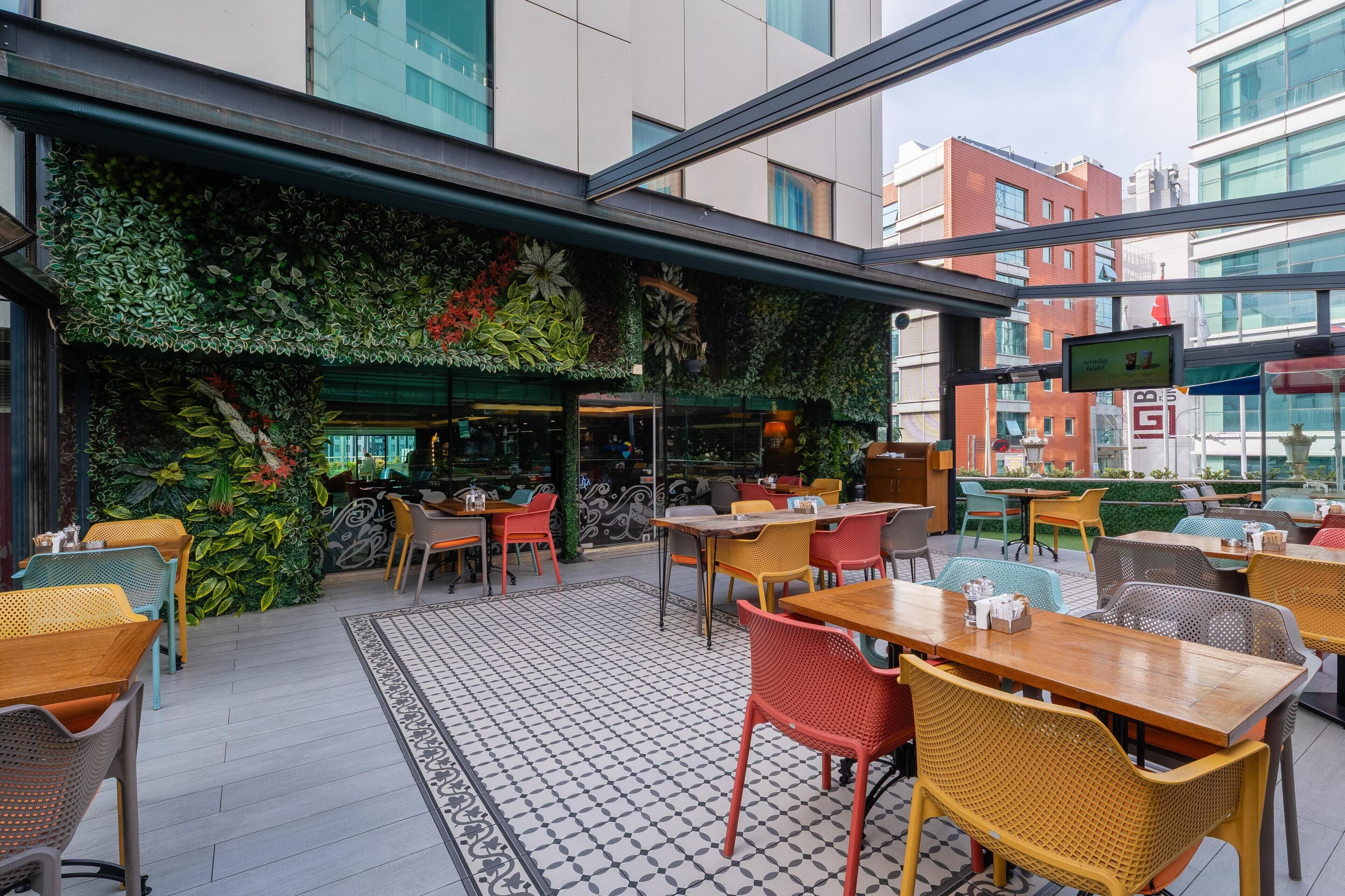 An outdoor seating area with tables and chairs, a patterned floor, and a green wall with plants.