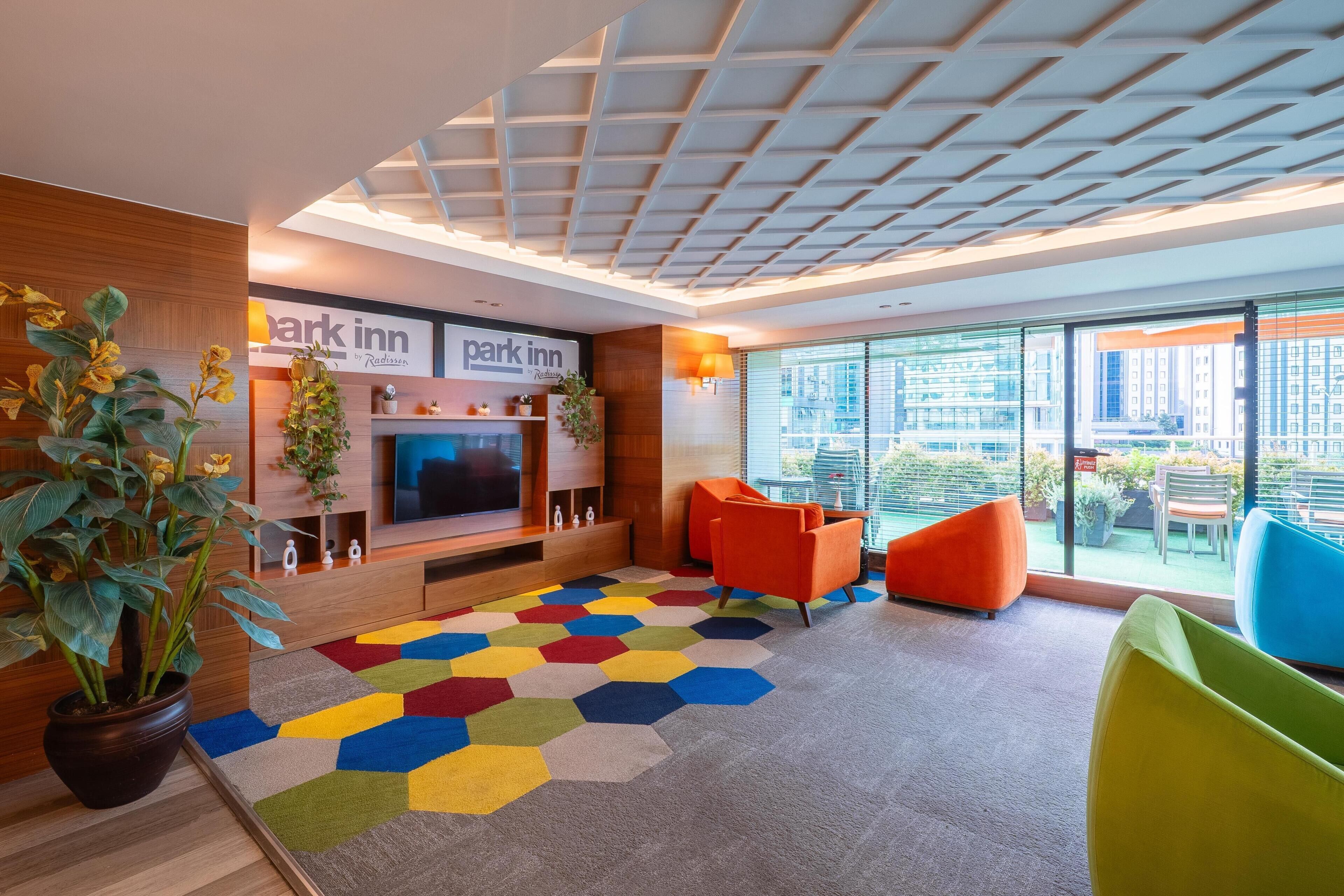 A modern hotel lobby with a large TV, colorful hexagonal carpet, and a view of the city through large windows.