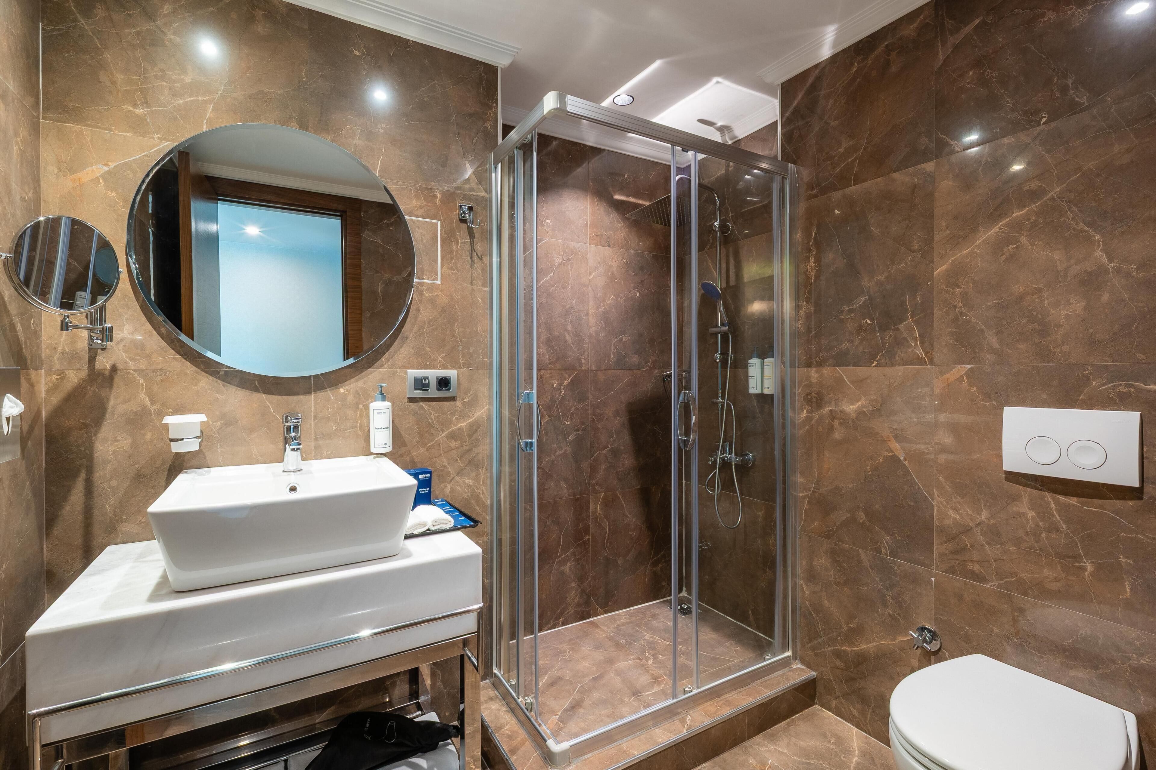 A modern bathroom with a glass-enclosed shower, a round mirror, and a white sink.