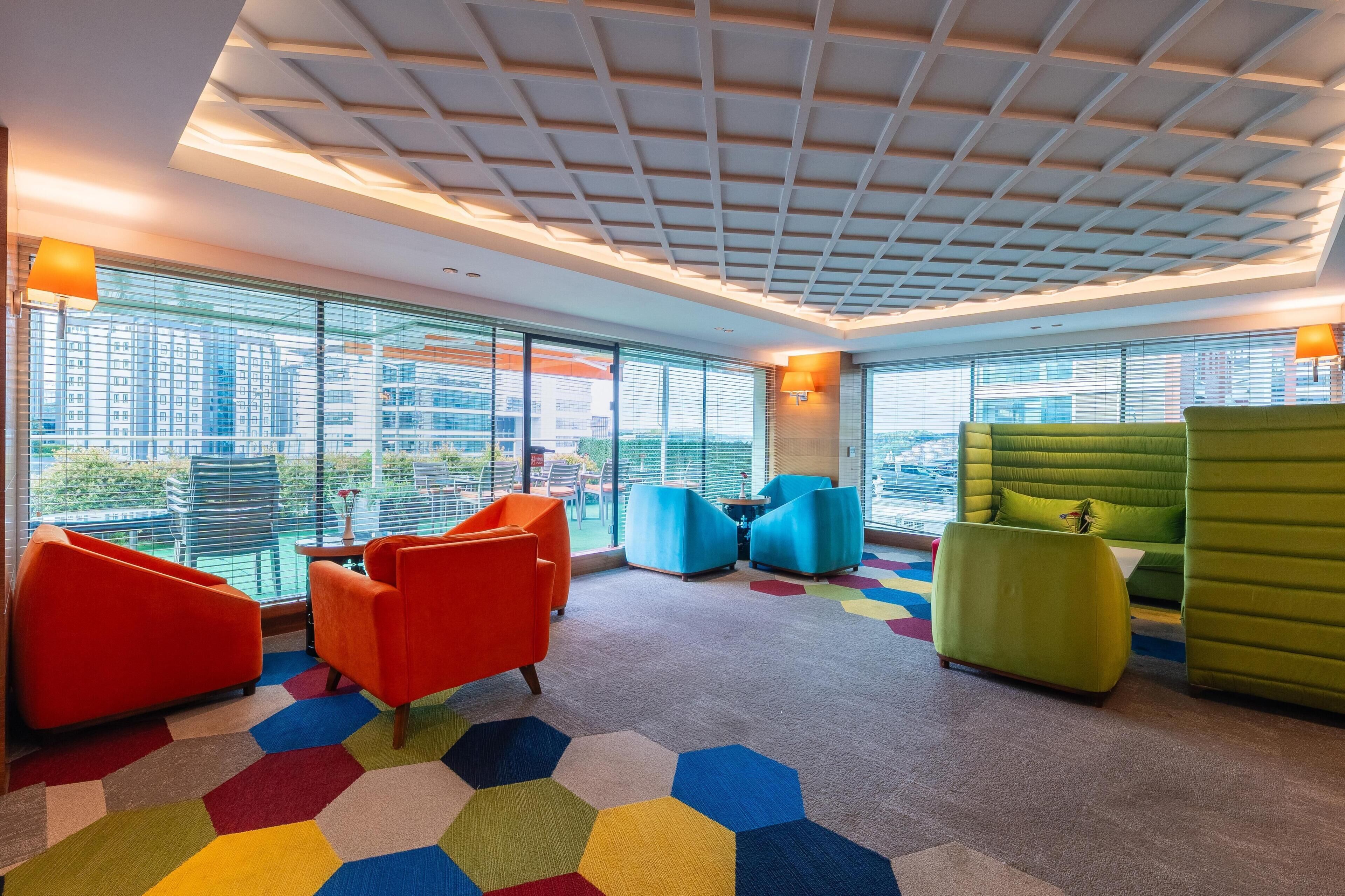 Un área de descanso moderna con alfombra hexagonal de colores, mobiliario contemporáneo y grandes ventanales con vistas a la ciudad.