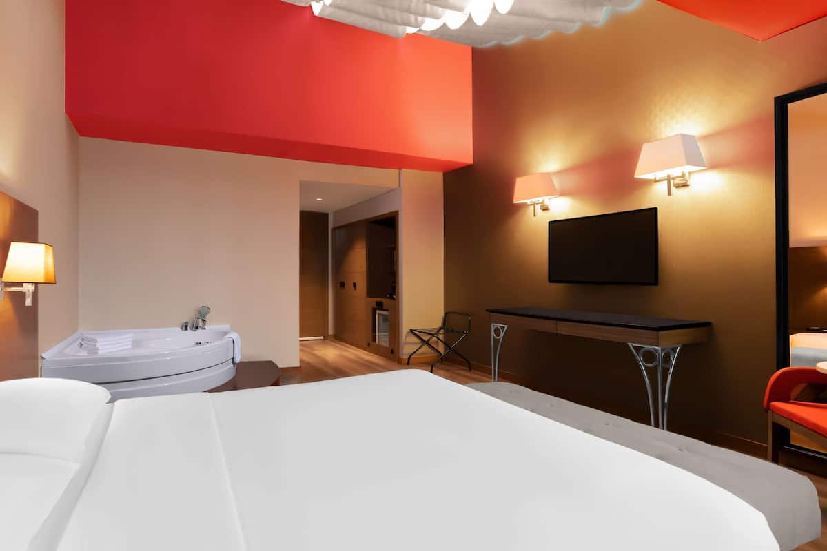 Ein Hotelzimmer mit einem großen Bett, einer Badewanne, einem Schreibtisch mit Stuhl und einem an der Wand befestigten Fernseher.