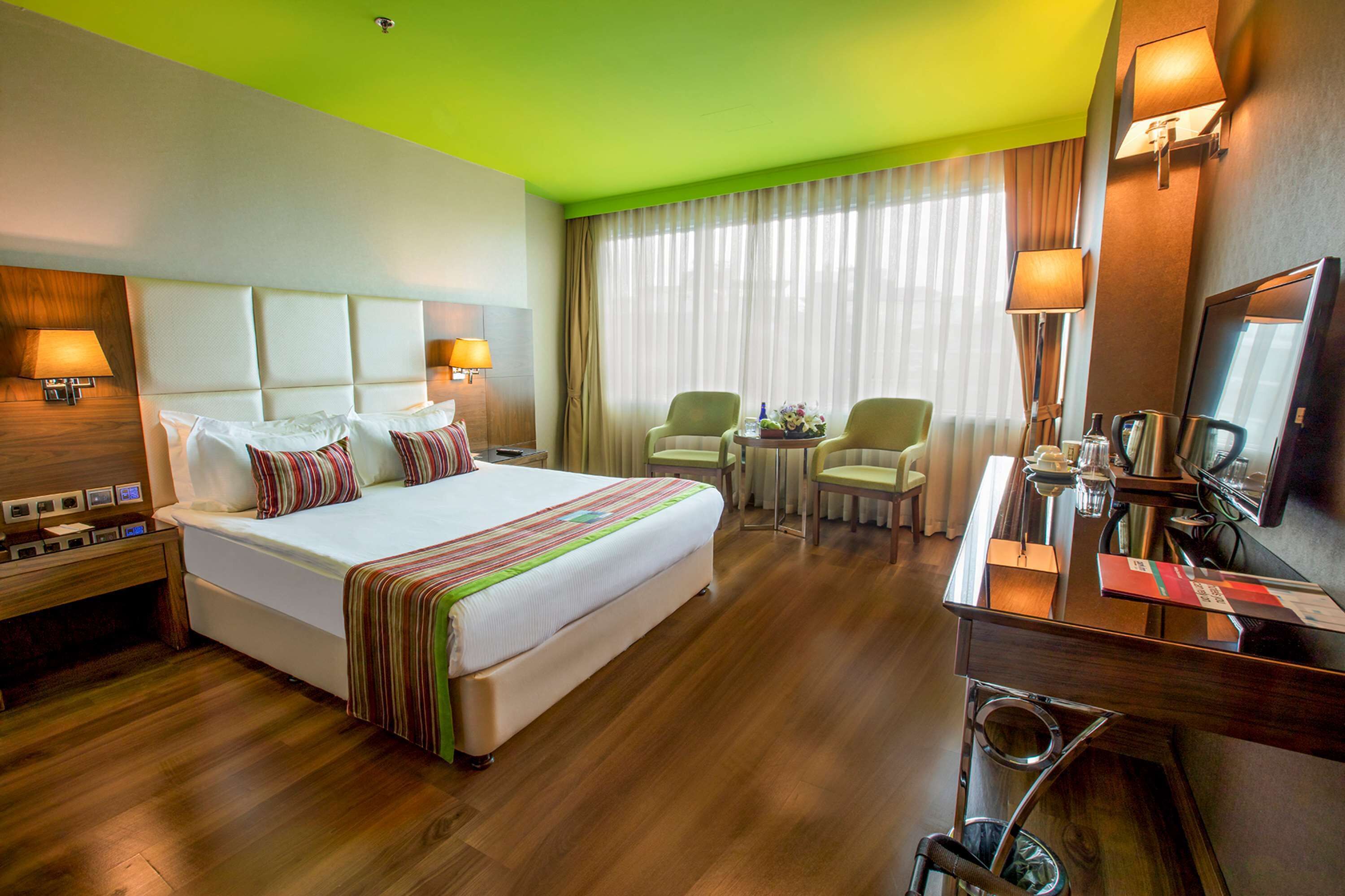 A hotel room with a large bed, two bedside tables, a desk with a chair, a TV, and a window with curtains.