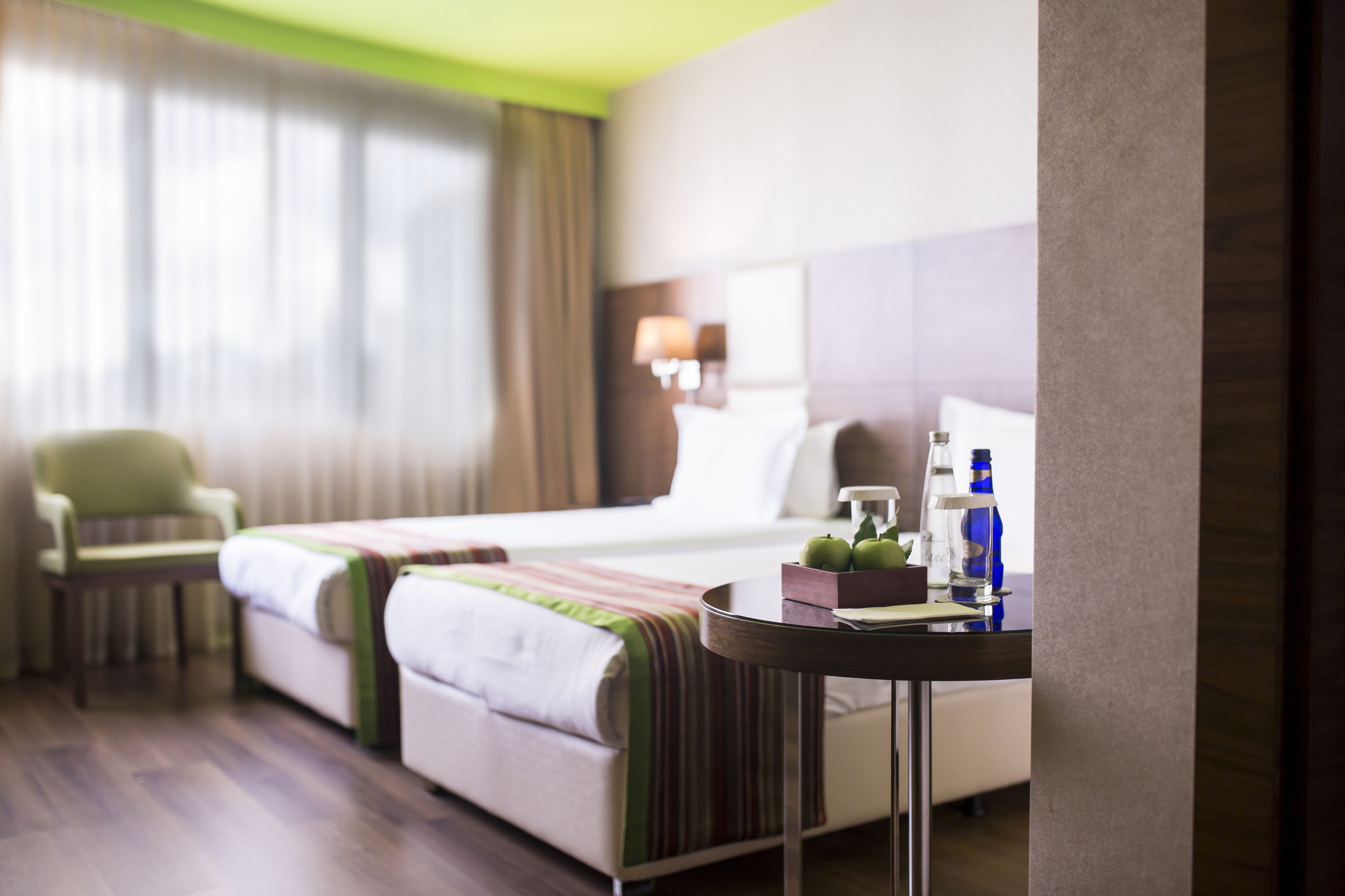 A hotel room with a bed, a chair, a table with a bottle, and a lamp.