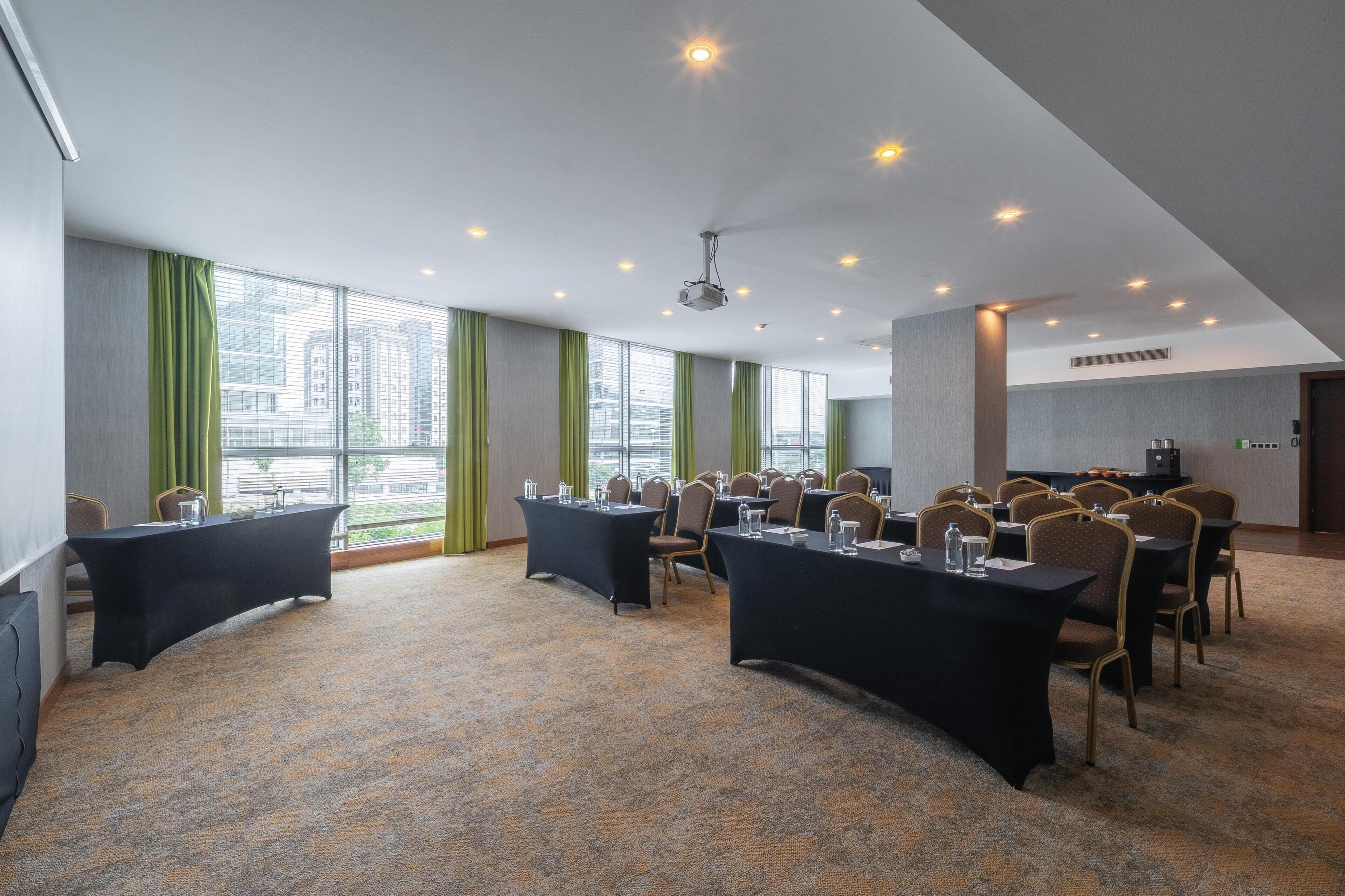 Une salle de conférence équipée de tables et de chaises, de grandes fenêtres avec des rideaux et d’un projecteur fixé au plafond.