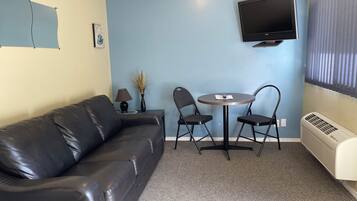 A room with a black leather sofa, a small round table with two chairs, a TV mounted on the wall, and a window with blinds.