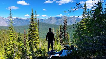 A person standing on a mountain trail with a view of a forest and mountains in the background.