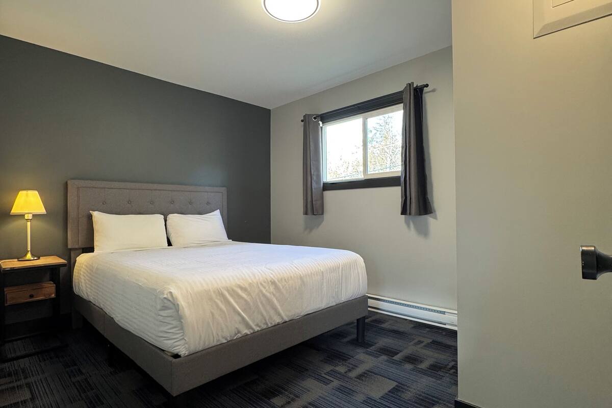 A bedroom with a bed, a nightstand, a lamp, a window with curtains, and a door.