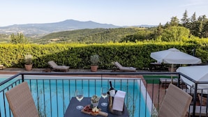 Zona de piscina con sillones de descanso, una mesa con copas de vino y una botella, y vistas a un paisaje montañoso.
