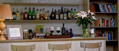 A bar area with a wooden counter, two chairs, a lamp, a vase with flowers, and a shelf with various bottles and books.