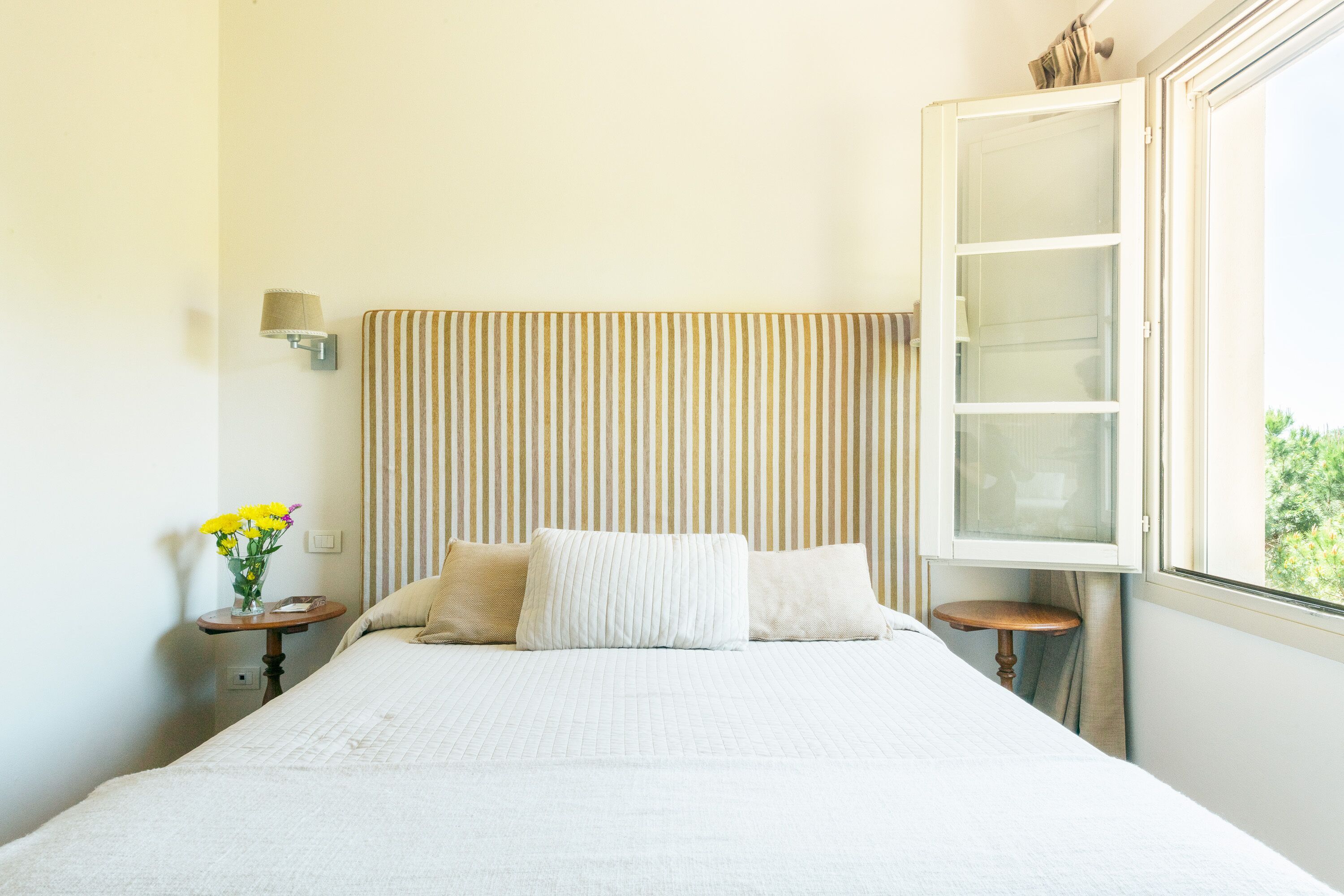 A neatly made bed with a striped headboard, a bedside table with a vase of flowers, and a window with a view of greenery.