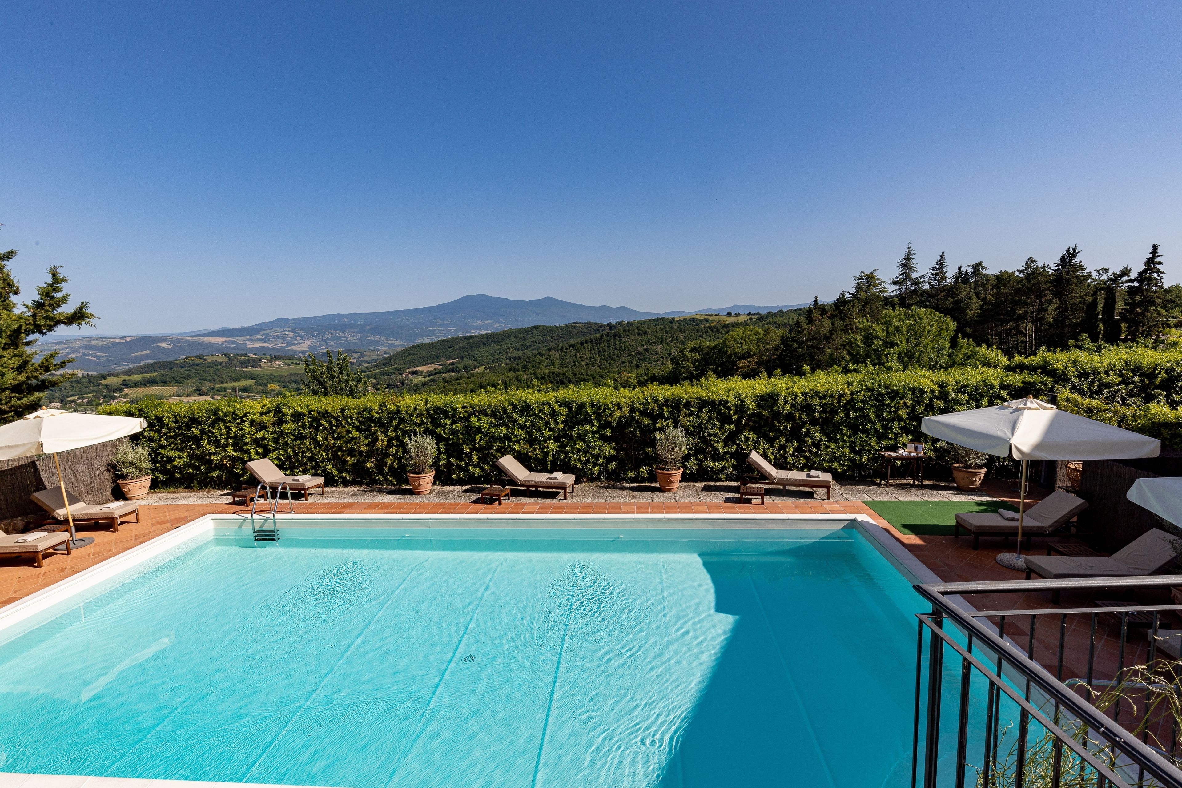 A rectangular swimming pool with clear blue water, surrounded by lounge chairs and umbrellas, set against a backdrop of green hills and a clear sky.