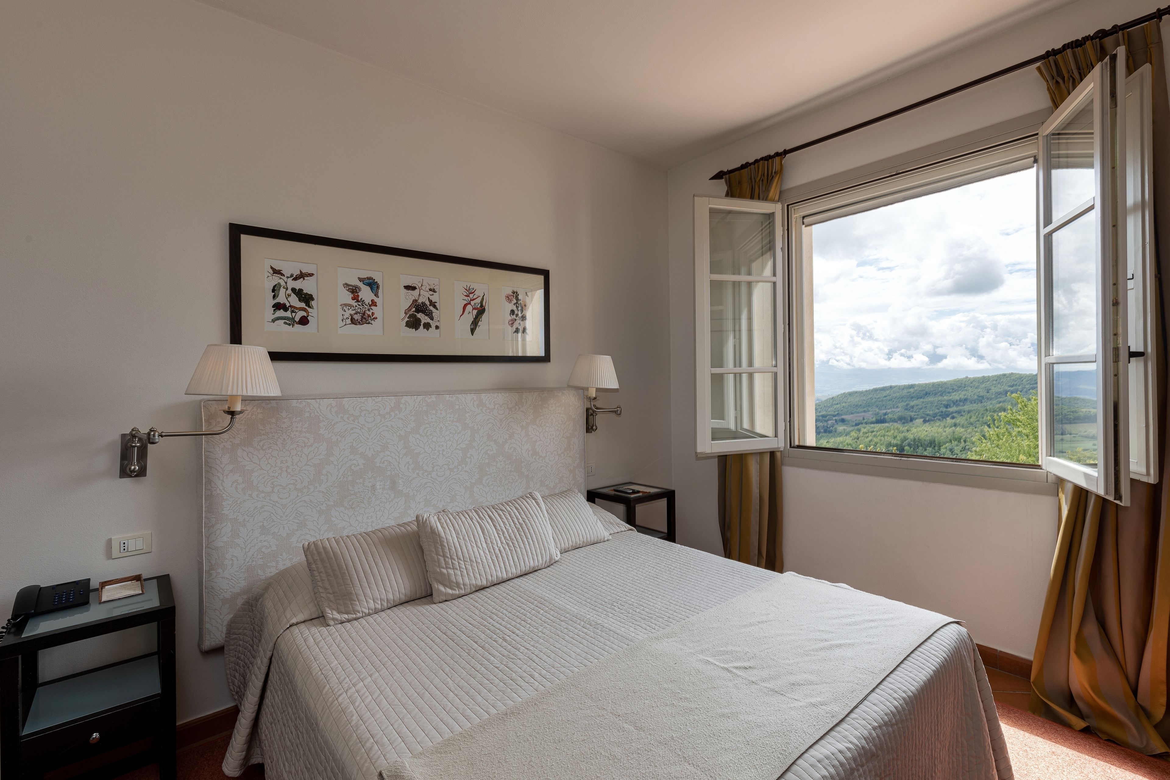 Un dormitorio con una cama, dos mesitas de noche, una lámpara de noche, una ventana con cortinas e impresión enmarcadas de motivos botánicos en la pared.