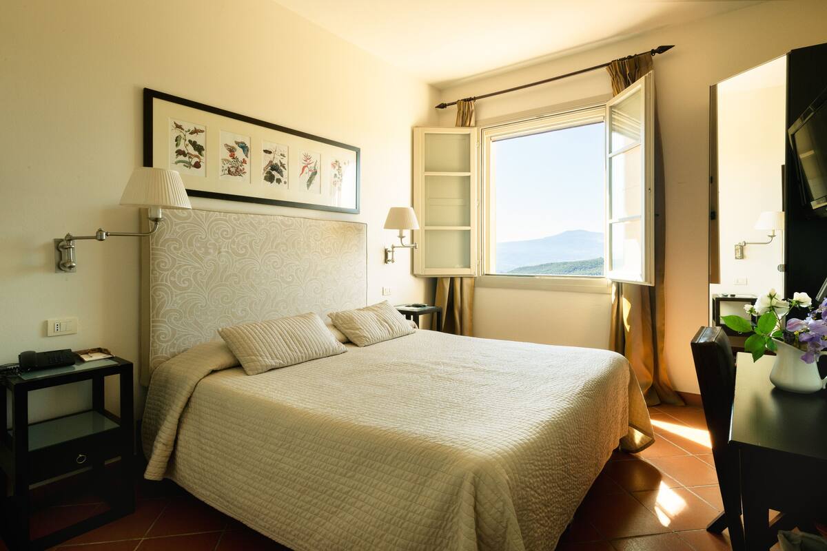 A bedroom with a bed, a nightstand, a lamp, a window with curtains, and a view of the mountains.