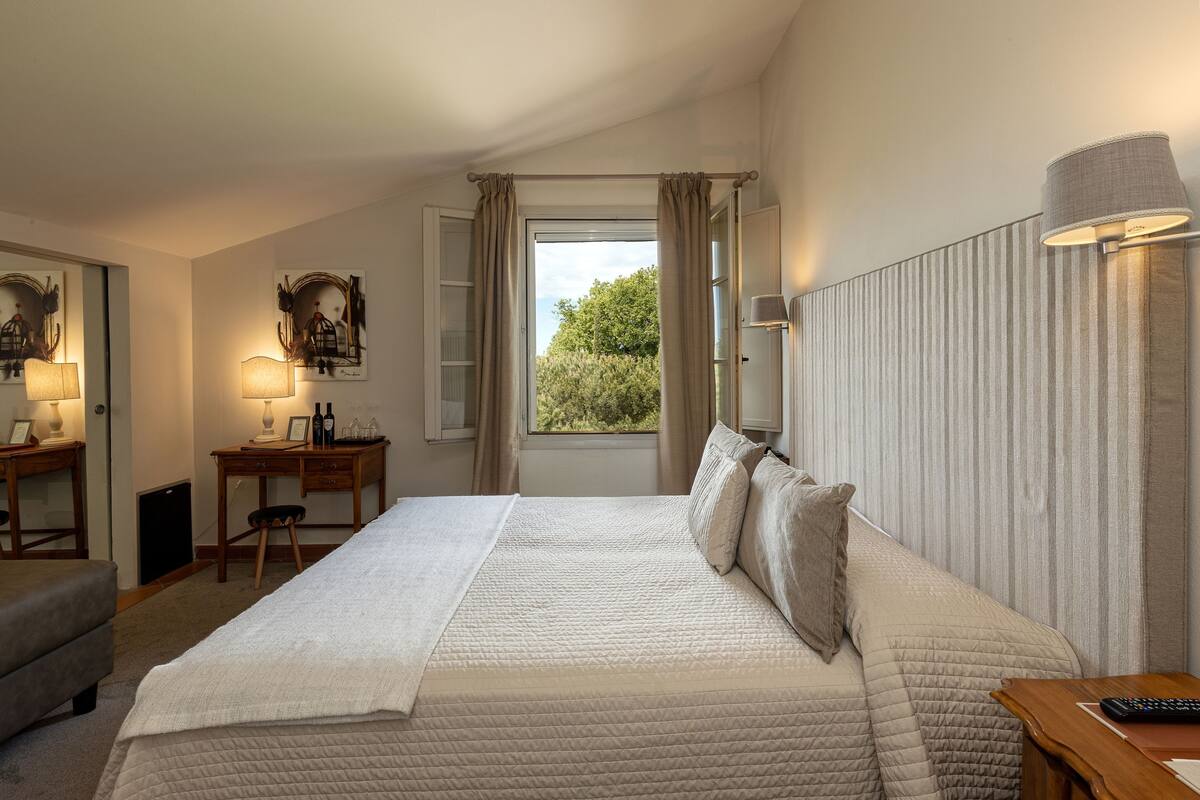 A bedroom with a large bed, a desk, a chair, a lamp, and a window with curtains.
