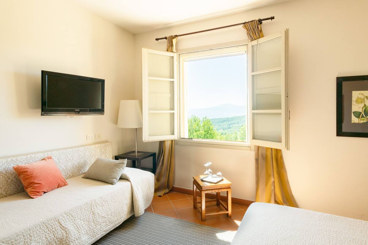 A hotel room with a bed, a sofa, a TV mounted on the wall, a small table with a lamp, and a window with curtains.