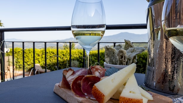 Una mesa con una copa de vino blanco, una jarra de metal y una tabla de madera con quesos y embutidos.