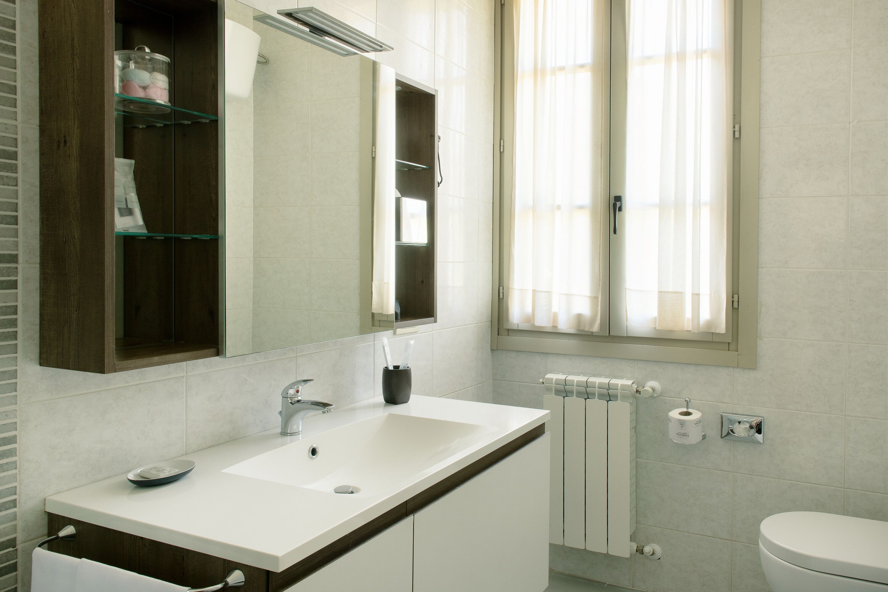 A modern bathroom with a white sink, mirror, and toilet. There is a window with curtains, a towel rack, and a shelf with toiletries.