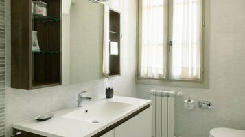 A modern bathroom with a white sink, mirror, and toilet. There is a window with curtains, a towel rack, and a shelf with toiletries.