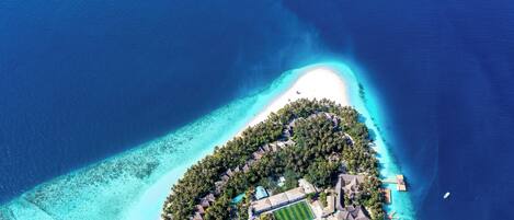 An aerial view of a tropical island resort with a central building surrounded by lush greenery, a sandy beach, and clear blue waters.