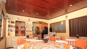 A dining area with tables covered in orange tablecloths and white chairs, a wooden ceiling, and a bookshelf with books and decorative items. - Canto do Mar (Ipojuca)
