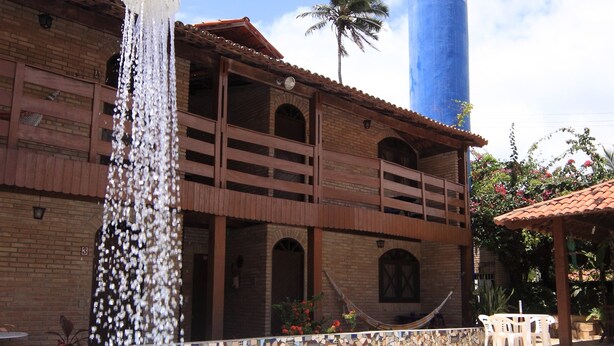 A swimming pool with a water fountain, a brick building with balconies, and a tall blue water tower. - Canto do Mar (Ipojuca)