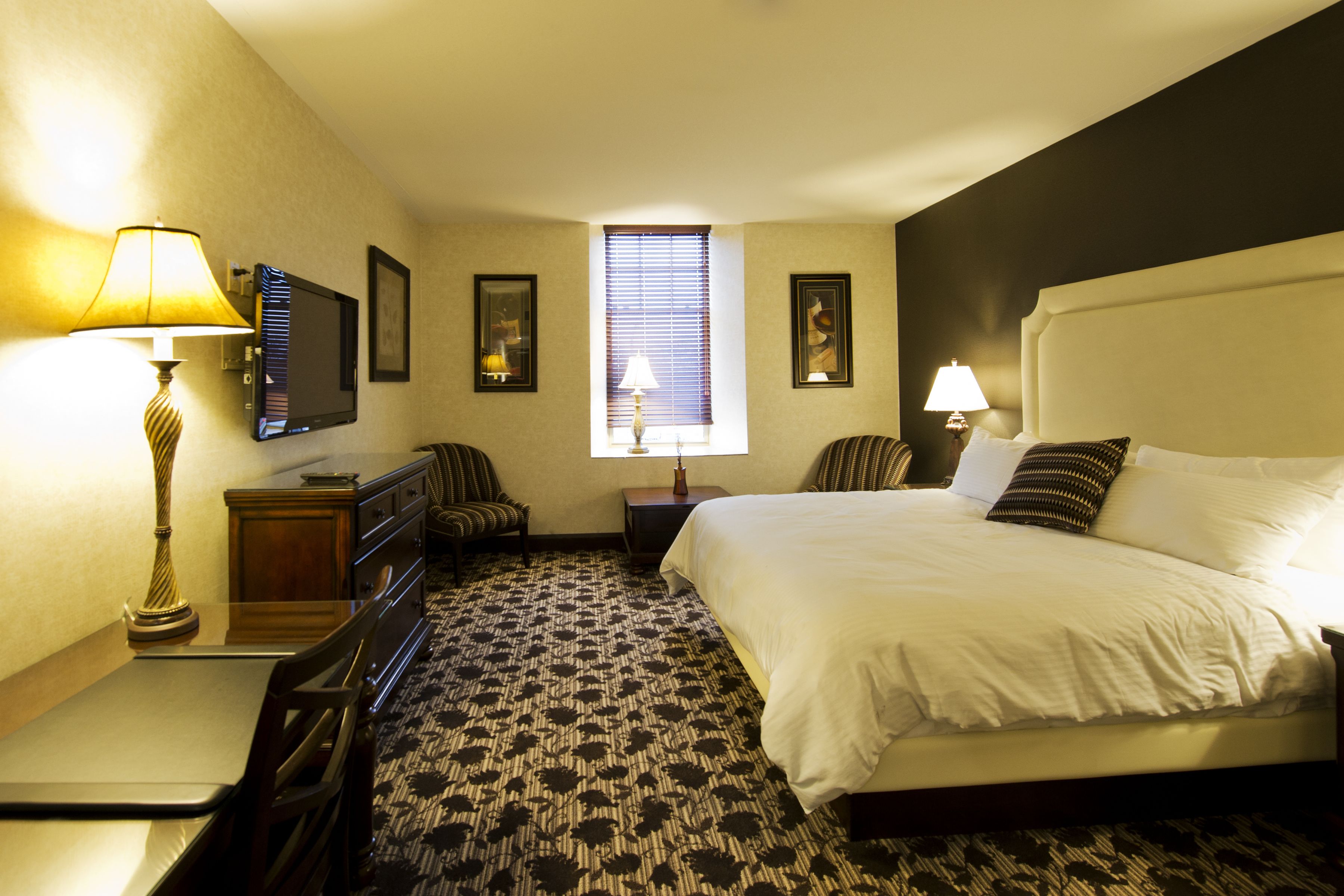A hotel room with a bed, a desk, a chair, a dresser, a TV, and a window with blinds.