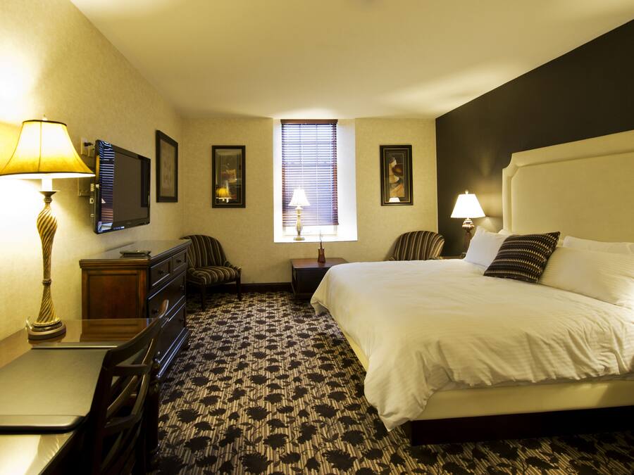 A hotel room with a bed, a desk, a chair, a dresser, a TV, and a window with blinds.