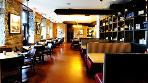 Restaurante com paredes de pedra, mesas e cabines de madeira. Quadros emoldurados adornam as paredes, e uma estante exibe diversos alimentos.