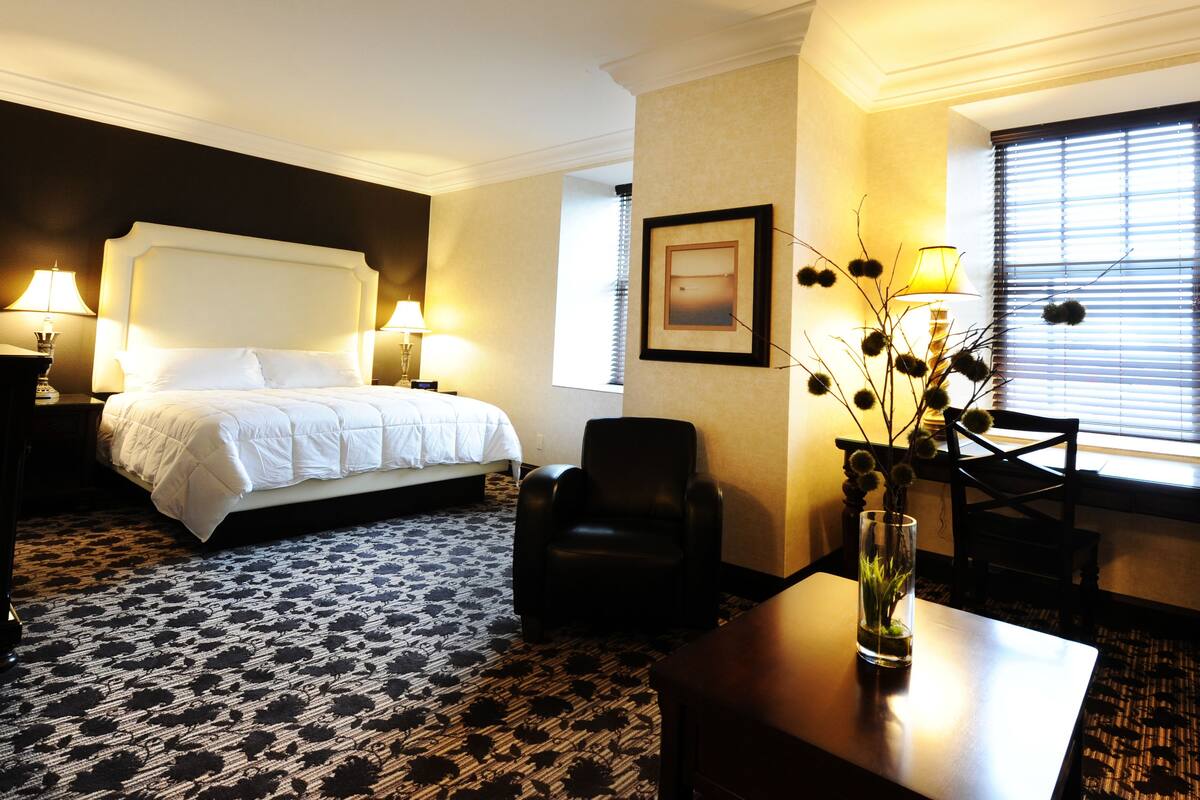 Una habitación de hotel con una cama grande, dos lámparas de noche, una silla, un escritorio, un florero con flores, un cuadro en la pared y una ventana con persianas.