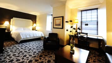 Ein Hotelzimmer mit einem großen Bett, zwei Nachttischlampen, einem Sessel, einem Schreibtisch, einer Vase mit Blumen, einem gerahmten Bild an der Wand und einem Fenster mit Rollläden.