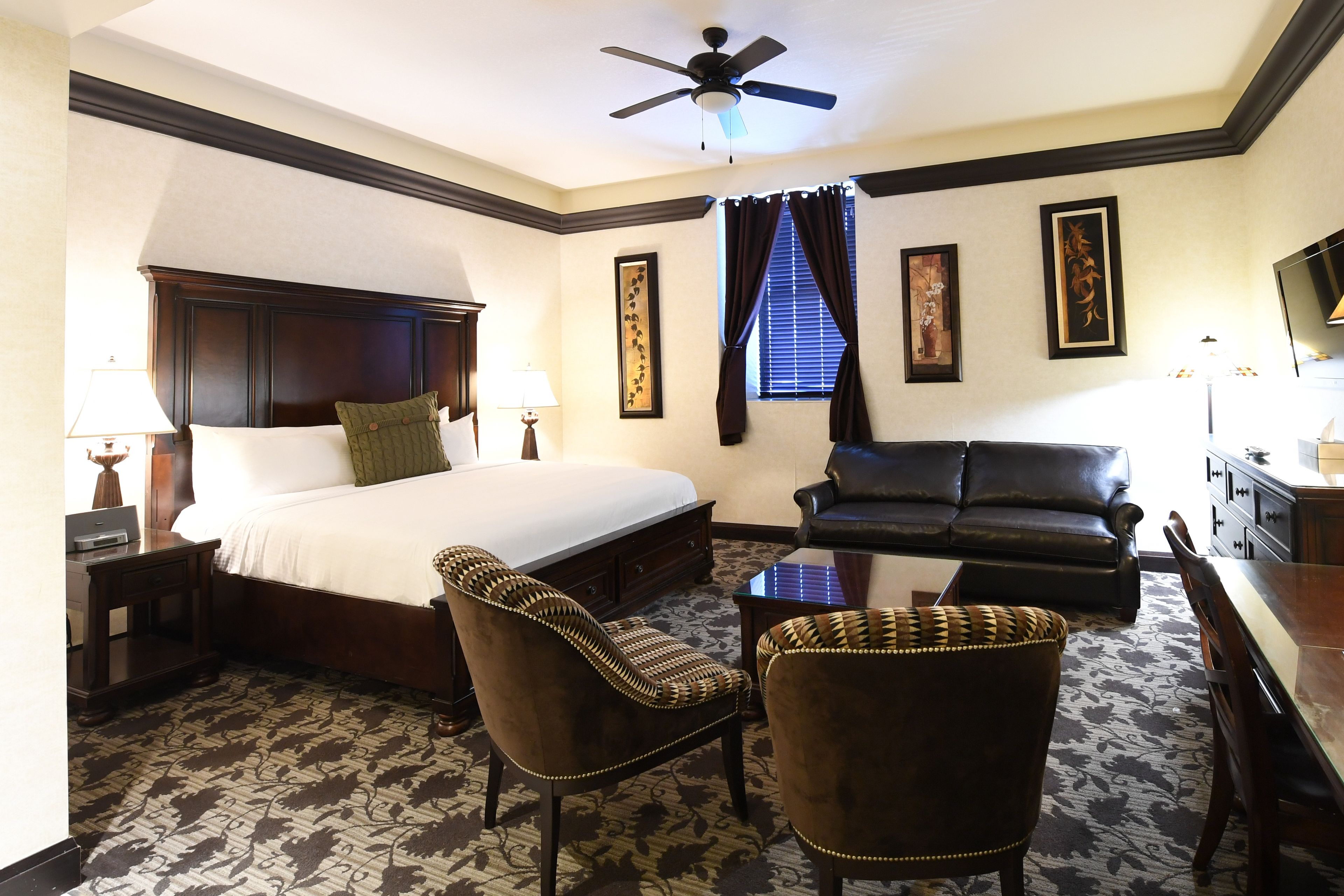 A hotel room with a large bed, a sofa, two armchairs, a desk, and a ceiling fan.