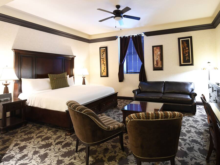 A hotel room with a large bed, a sofa, two armchairs, a desk, and a ceiling fan.