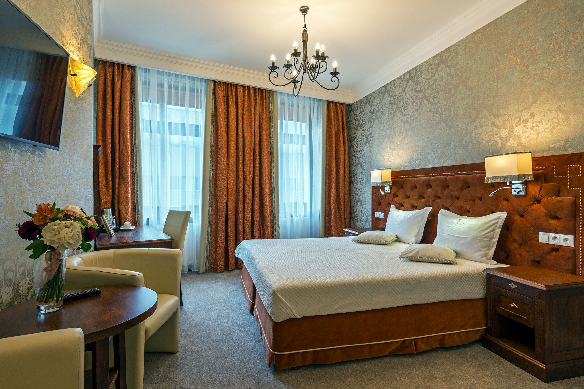 A hotel room with a large bed, a desk with a chair, a television, and a chandelier.