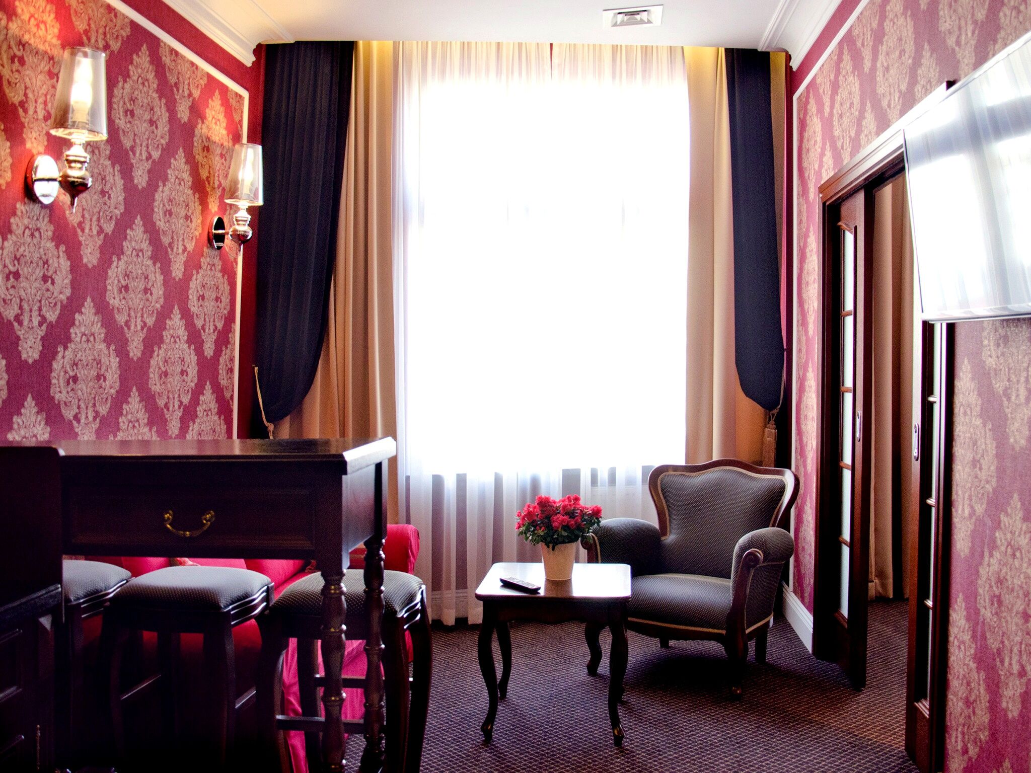 A hotel room with a desk, two chairs, a small table with a vase of flowers, and a large window with curtains.