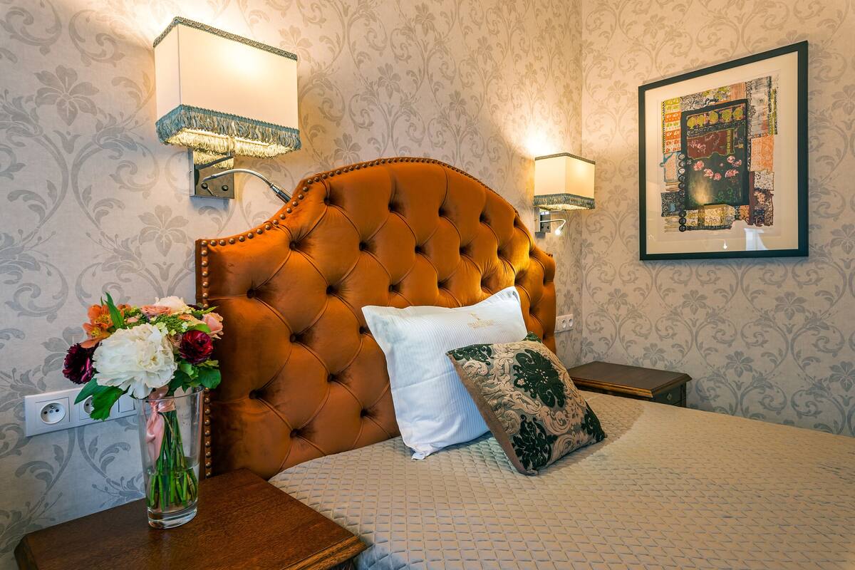 A hotel room with a tufted headboard, a vase of flowers, a bed with patterned bedding, and wall-mounted lamps.