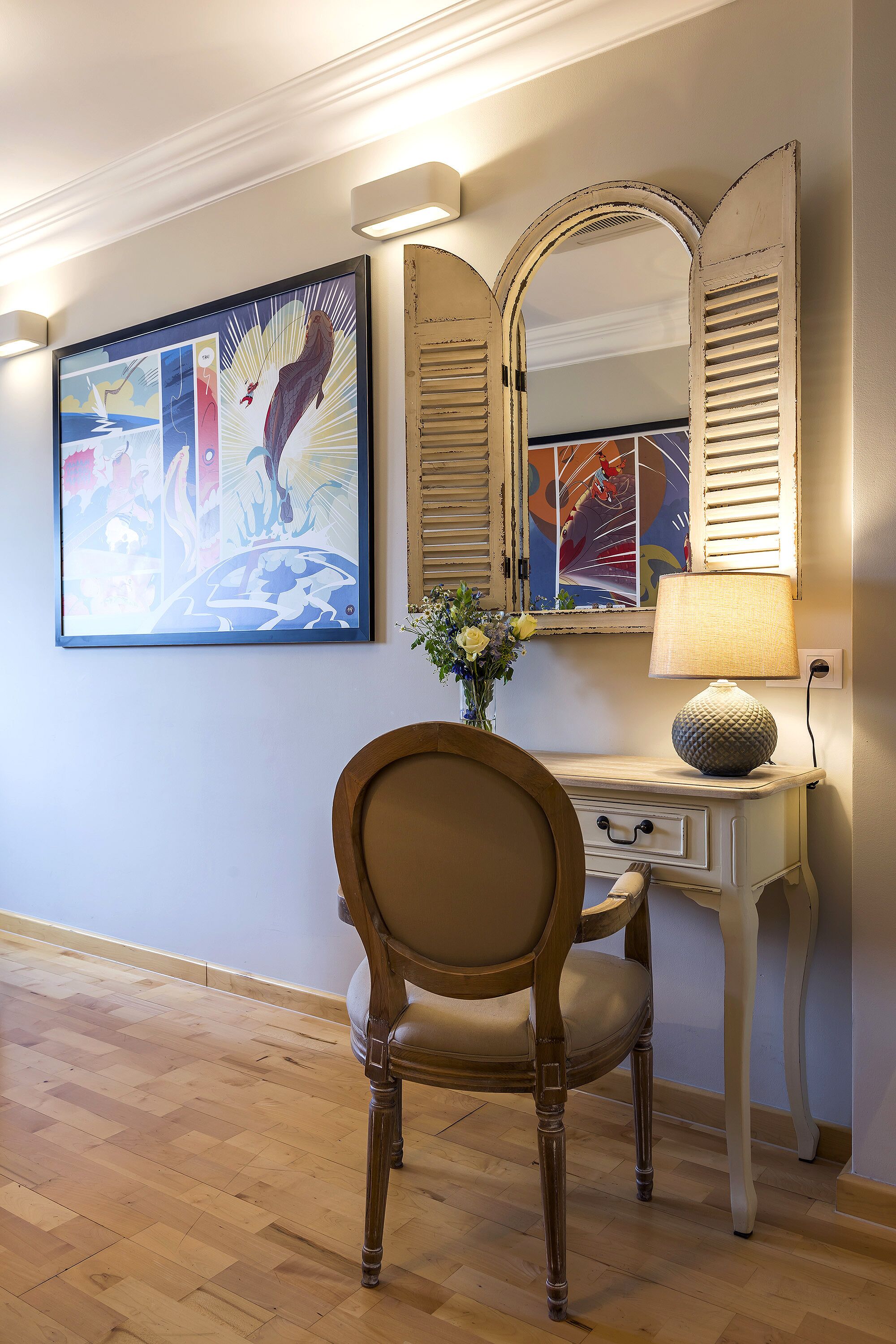 A room with a wooden floor, a chair, a desk with a lamp, and a wall with a framed picture and a mirror.