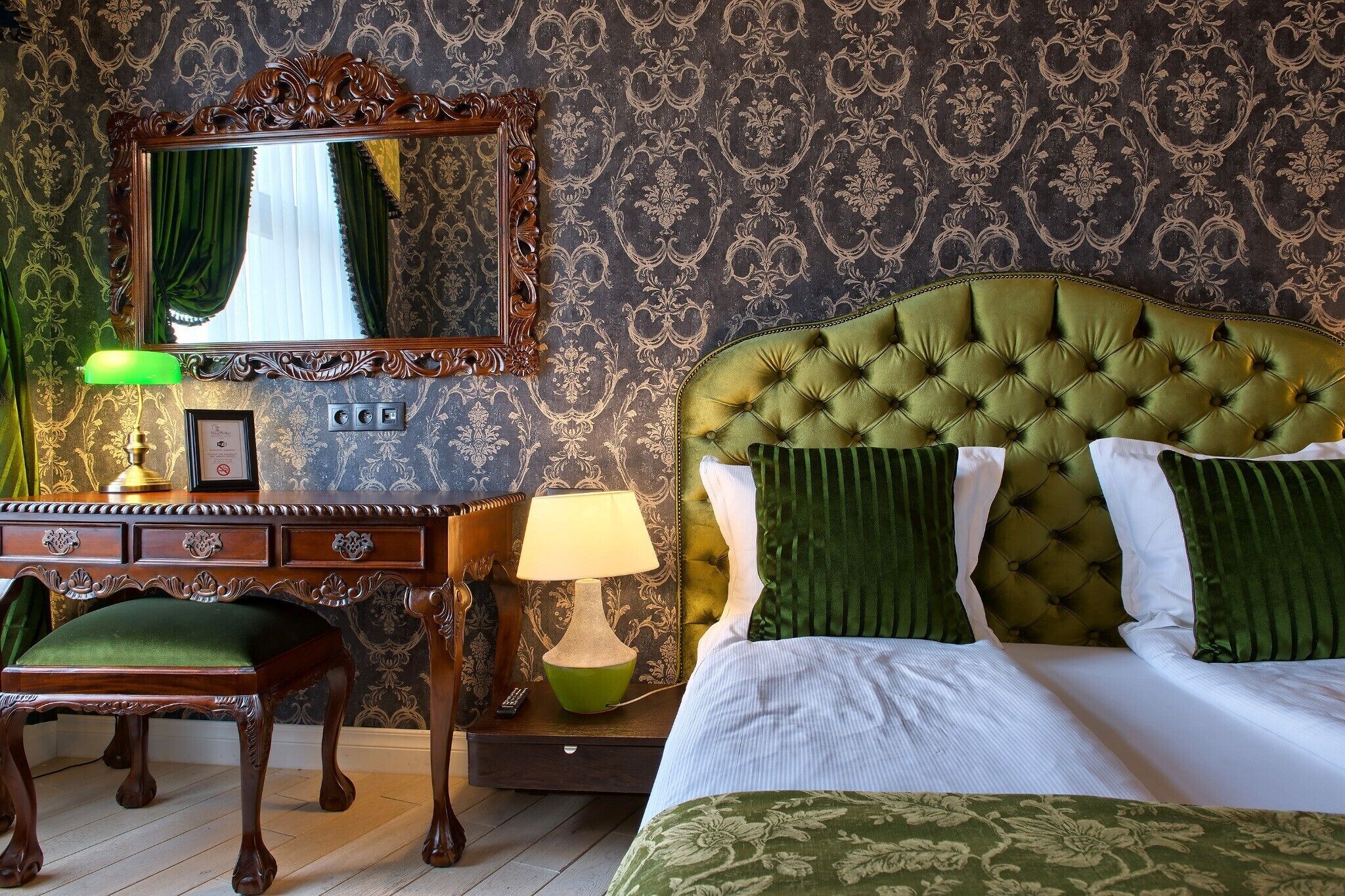 Una habitación con un cabecero tapizado en verde, un escritorio de madera con una silla verde, un espejo con un marco decorativo y una lámpara verde.