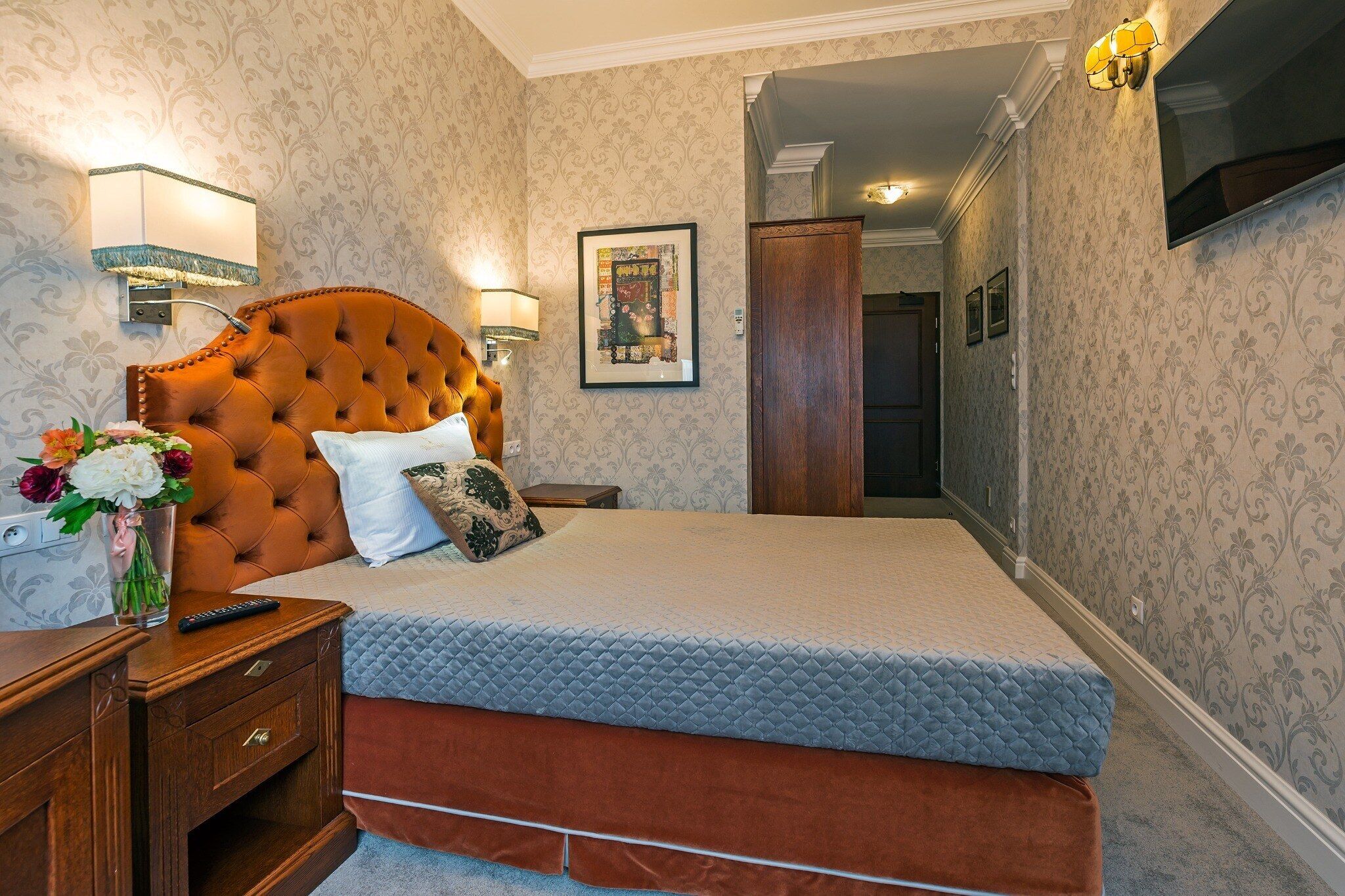Quarto de hotel com cama grande, mesa de cabeceira de madeira, vaso com flores, quadro na parede e televisão fixada na parede.