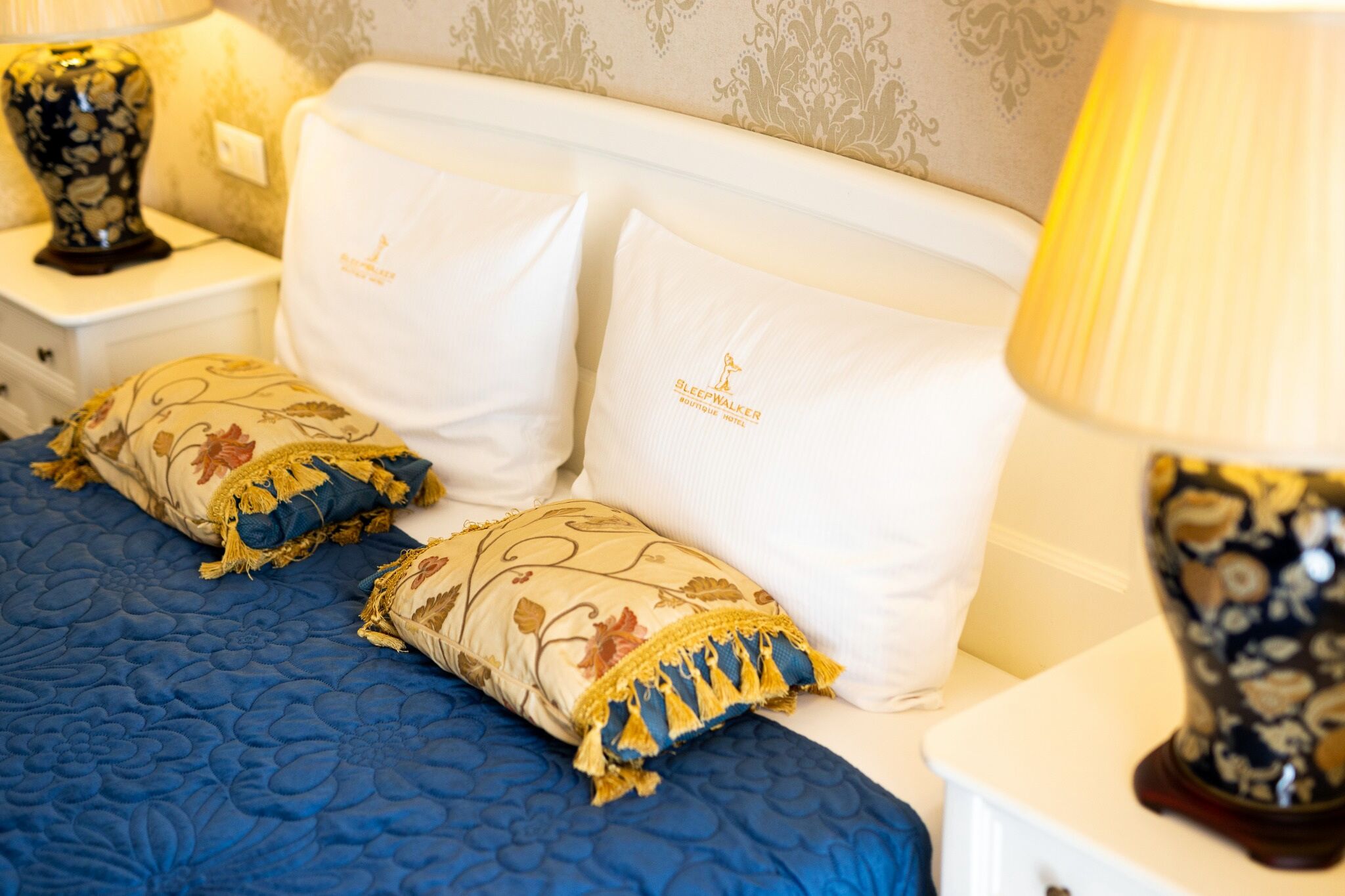 A bed with blue bedding and decorative pillows, a bedside lamp, and a nightstand with a vase.