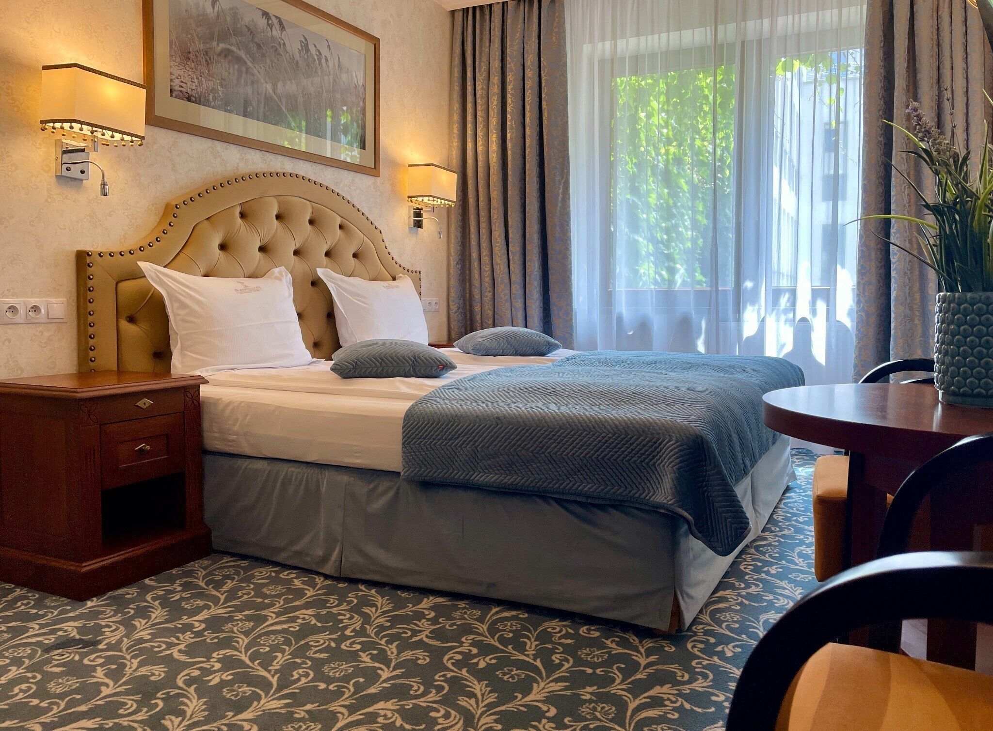 Ein Hotelzimmer mit einer gepolsterten Kopfteile, einem Bett mit blauer Decke, einem Nachttisch, einem Sessel und einem Fenster mit Vorhängen.