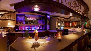 Zona de bar con mostrador de madera, estantes llenos de botellas y una lámpara decorativa.