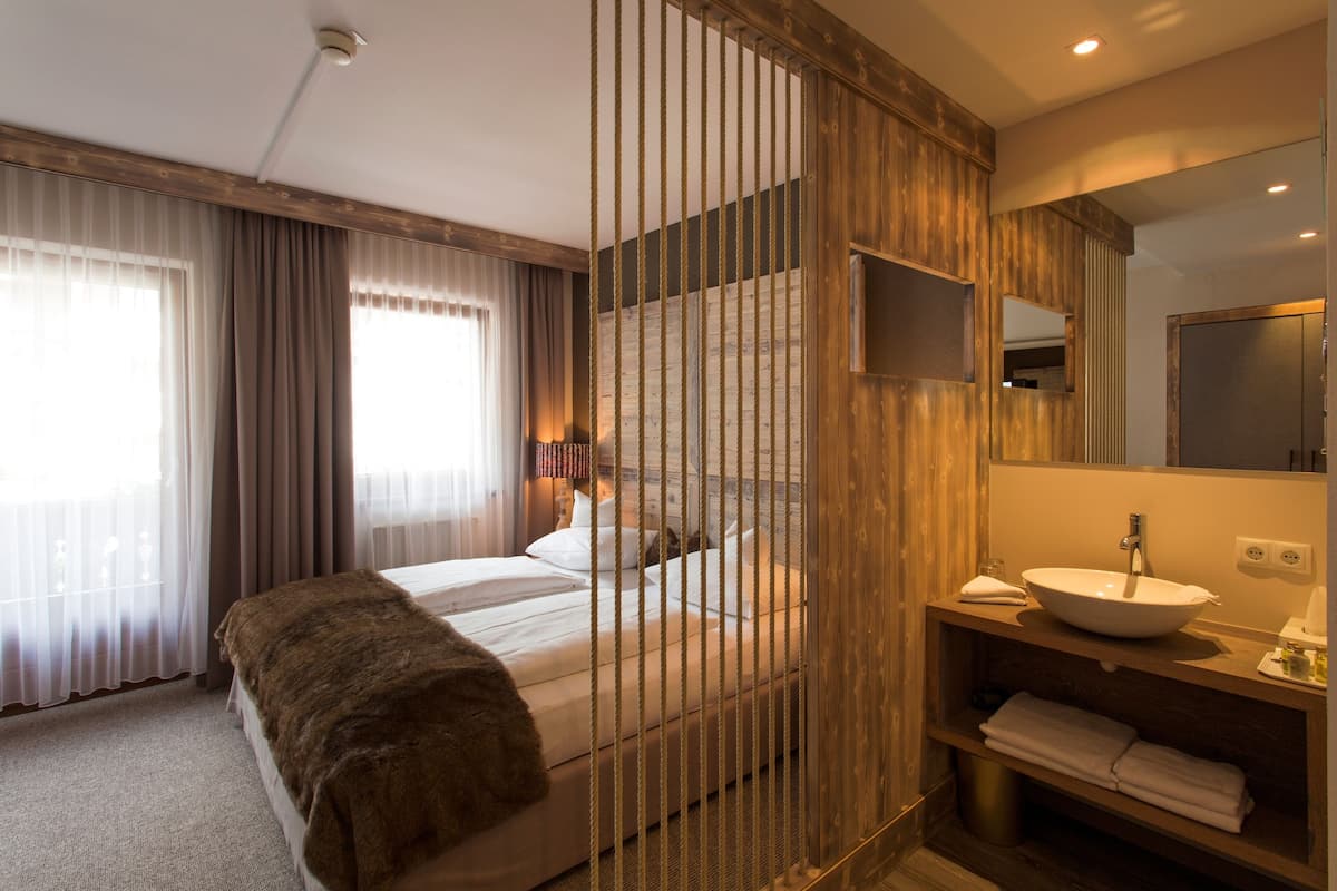 Una habitación de hotel moderna con una cama, un separador de madera, un baño con lavabo y espejo, y una ventana con cortinas.